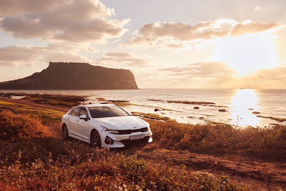 一台白色現代的車，行駛在濟州島海岸線上，後方是城山日出峰